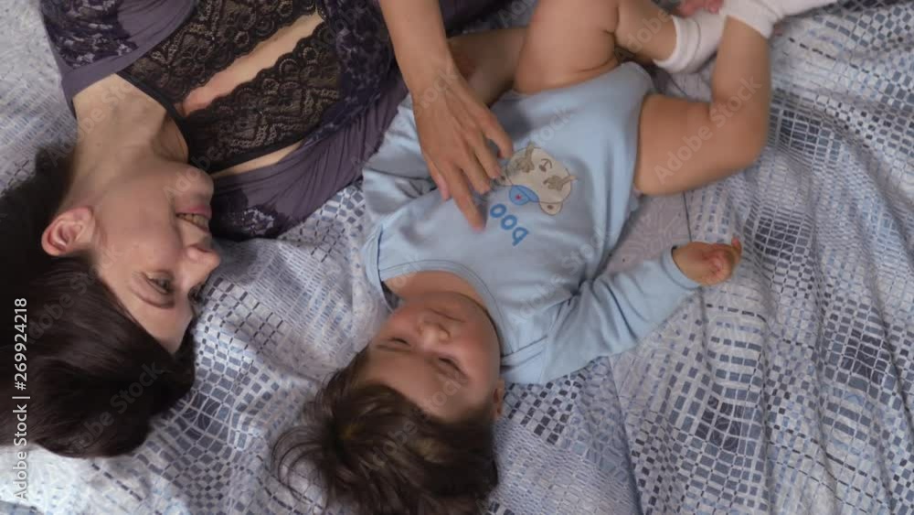 Pregnant Mother laying with her baby son on the bedroom bed playing and having fun - Asian mixed ethnicity child Boy wearing blue body shirt with smiling bear cartoon
