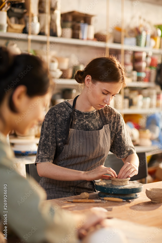 Serious concentrated young female potter in gray stripped apron sitting at desk and moulding clay bowl with hands before firing