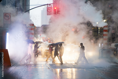 Undefined people with umbrellas are crossing the 42nd street in Manhattan. Steam coming out from from the manholes in the background. Manhattan, New York City, Usa. photo