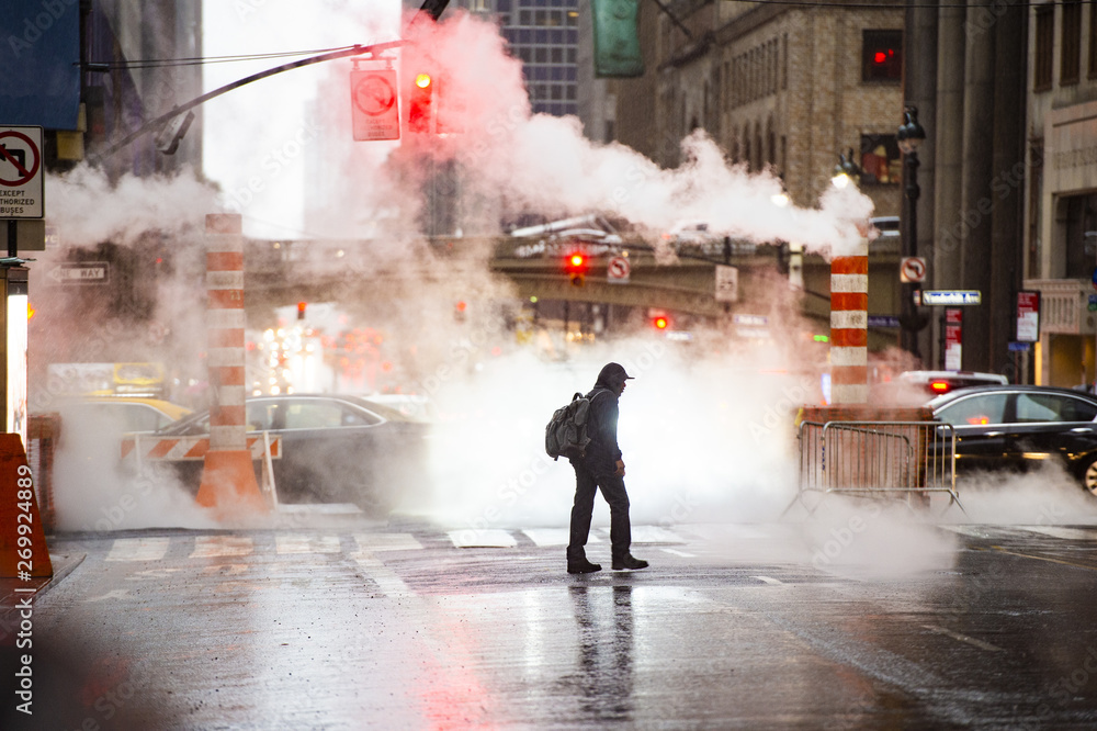 A black male is crossing the 42nd street in Manhattan while steam coming out from from the manholes. 42nd Street is a major crosstown street in Manhattan, New York, USA.