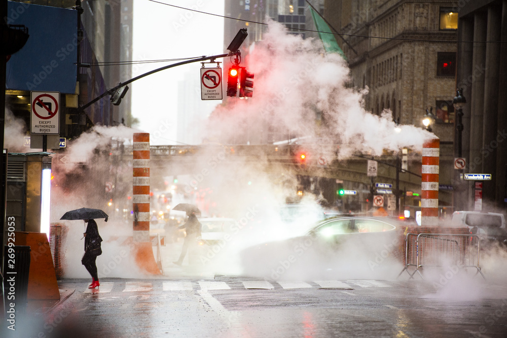 A woman with an umbrella and red high heels shoes is crossing the 42nd street in Manhattan. Cars and steam coming out from from the manholes in the background. New York City, Usa.