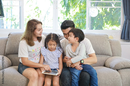 Happy Asian family teaching their childrens son and daughter how to use tablet while sitting on grey sofa in living room with smiling faces