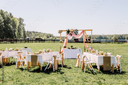 The chairs and table for guests, decorated with candle and floral arrangement, served with cutlery, crockery. Wedding banquet on a green lawn in the field, on the territory of the stable. Deer farm.