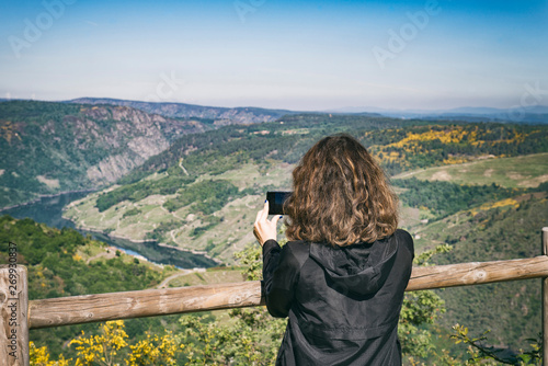 A woman in a dark jacket and hood is taking photos with her mobile on a sunny day in a beautiful viewpoint of the Ribeira Sacra