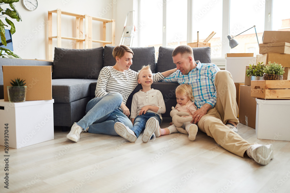 Group of positive young family in casual outfits sitting on floor and leaning on sofa while getting settled in new home Group of positive young family in casual outfits sitting on floor and leaning on sofa while getting settled in new home