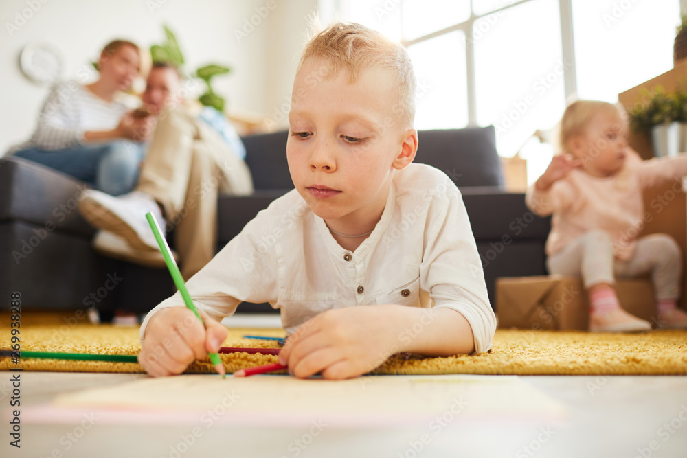 Serious blond-haired boy in casual sweater lying on soft carpet and using colorful pencil while drawing picture at home Serious blond-haired boy in casual sweater lying on soft carpet and using colorful pencil while drawing picture at home