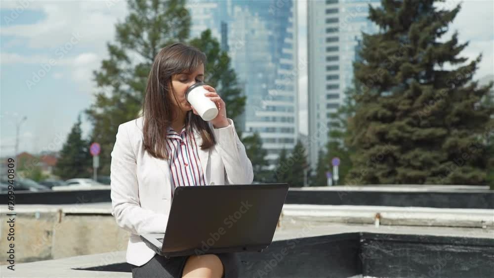 Businesswoman Using Laptop Outdoors while Having Breakfast. She is Drinking Coffee while Sitting at a Bench. Lifestyle and Business Concept