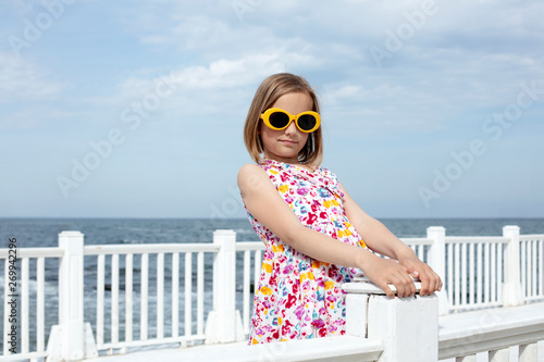 Small (8 years old) pretty cheerful girl in a dress with a floral print and in yellow sunglasses stands on a wooden white pier.