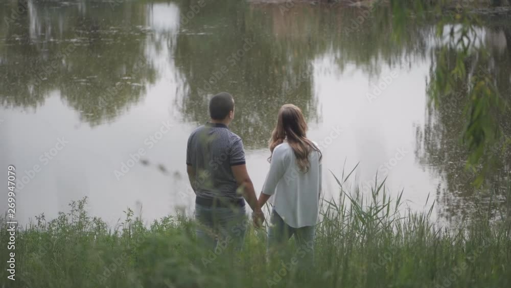 Young man and woman standing at the pond and look into the distance holding hands. Young man and woman standing at the pond and look into the distance holding hands.