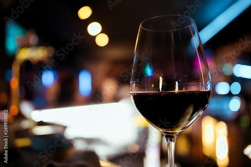Low angle close up perspective of crystal clear wine glass with traditional round goblet shape filled with dark red wine and slim stem on wood counter top bar with blurry restaurant background scene