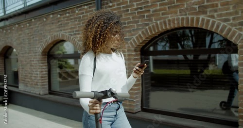 TRACKING attractive African American black female walking in the street with an electric scooter, checking her phone. 4K UHD 60 FPS SLOW MOTION photo