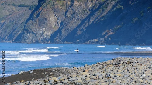 Surfer standing at the shoreline ready to paddle out into California surf with large scenic cliffs in the background
