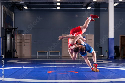 Two young men in blue and red wrestling tights are wrestlng and making a suplex wrestling on a yellow wrestling carpet in the gym. The concept of fair wrestling