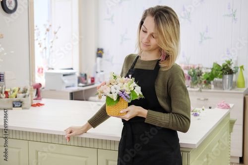 A woman florist in a black apron holds a floral composition in her hands. Young female flower seller smiling looking at bouquet.