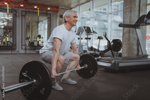 Full length shot of a senior sportsman working out with barbell at gym, copy space. Elderly weightlifter exercising with heavy barbell. Aged fitness man doing deadlift at sport studio