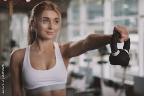 Selective focus on a kettlebell beautiful sportswoman is lifting. Attractive young fitness woman smiling, working out with kettlebell at the gym