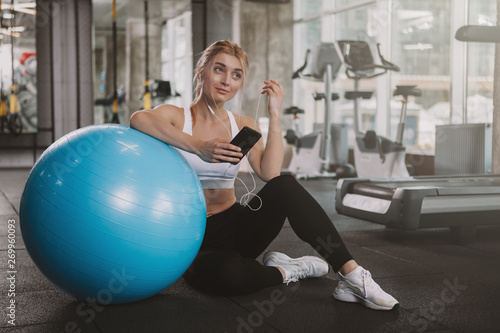 Gorgeous happy healthy young sportswoman using her smart phone at the gym, copy space. Lovely blond haired athletic woman relaxing after working out with fitness ball, copy space © Ihor
