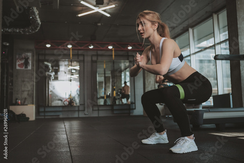 Athletic healthy blond haired woman squatting with rubber resistance band, copy space. Attractive strong sportswoman doing squats at the gym. Body care, healthy lifestyle concept