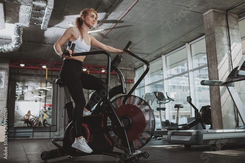 Healthy athletic woman doing intense workout on air bike at the gym, copy space. Fitness female with toned strong body cycling at sport studio, using air bicycle