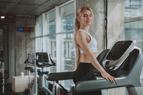 Happy healthy beautiful woman smiling to the camera, walking on a treadill at sport studio, copy space. Charming blond haired fitness female enjoying morning workout at the gym, doing cardio