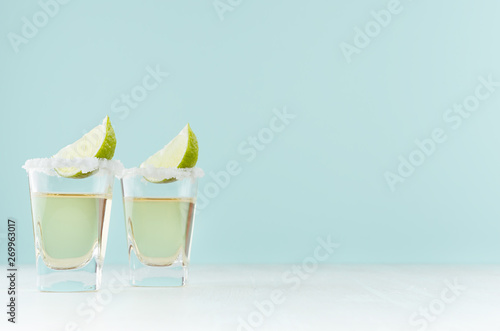 Photo Golden mexican tequila shot drinks with salty rim, piece lime in elegant glass on pastel green wall, white wood table, copy space