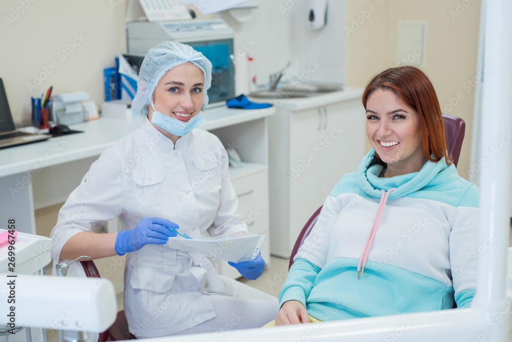 Young beautiful female dentist and patient happy are looking directly into the camera. Red-haired woman sits in a chair with an orthodontist and smiles. Dental services. Young beautiful female dentist and patient happy are looking directly into the camera. Red-haired woman sits in a chair with an orthodontist and smiles. Dental services.