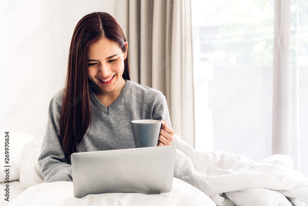 Young woman relaxing and drinking cup of hot coffee or tea using laptop computer on a cold winter day in the bedroom.woman checking social apps and working.Communication and technology concept Young woman relaxing and drinking cup of hot coffee or tea using laptop computer on a cold winter day in the bedroom.woman checking social apps and working.Communication and technology concept