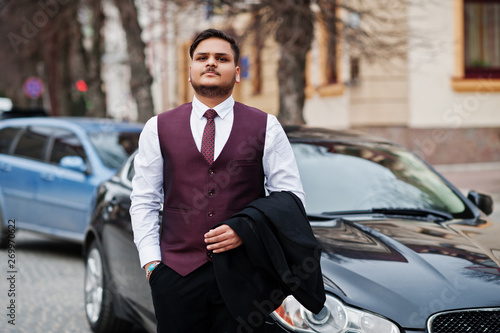 Stylish indian businessman in formal wear vest suit standing against black business car on street of city.