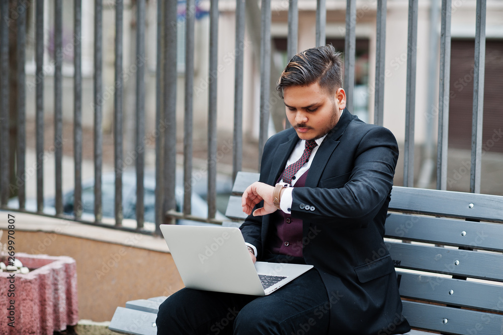 An important meeting. Stylish indian businessman in formal wear sitting on bench with laptop and looking at watches. An important meeting. Stylish indian businessman in formal wear sitting on bench with laptop and looking at watches.