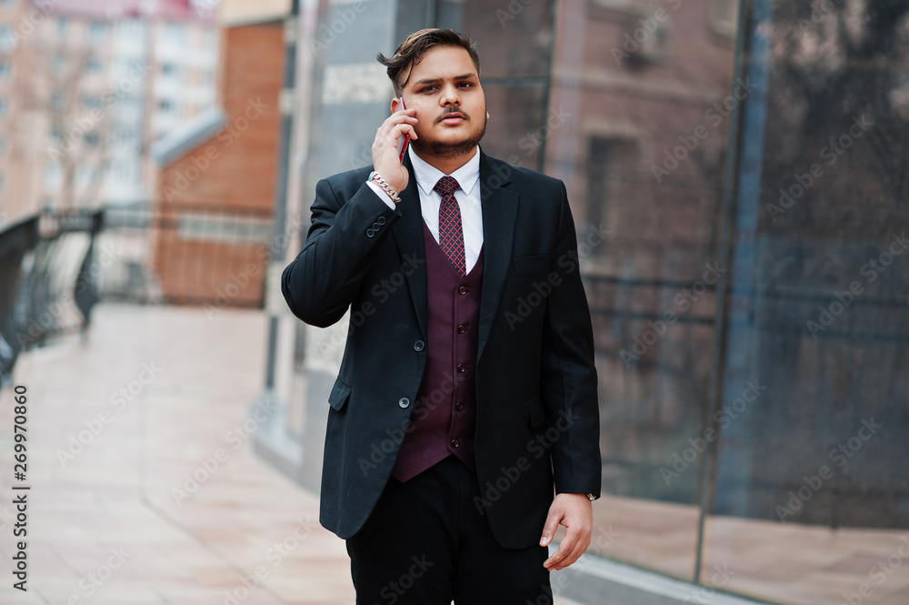 Stylish indian businessman in formal wear standing against windows in business center and speaking on phone. Stylish indian businessman in formal wear standing against windows in business center and speaking on phone.