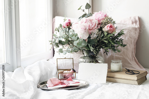 Spring, summer wedding still life scene. Blank paper card mockup, old books and linen pillow at windowsill. Vintage feminine floral composition. Bouquet of pink roses, peony flowers and eucalyptus. © tabitazn