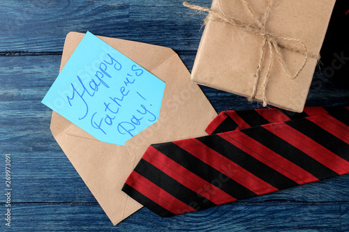 Happy father's day. Text on paper in an envelope, men's tie and gift on a blue wooden table. men's holiday. top view