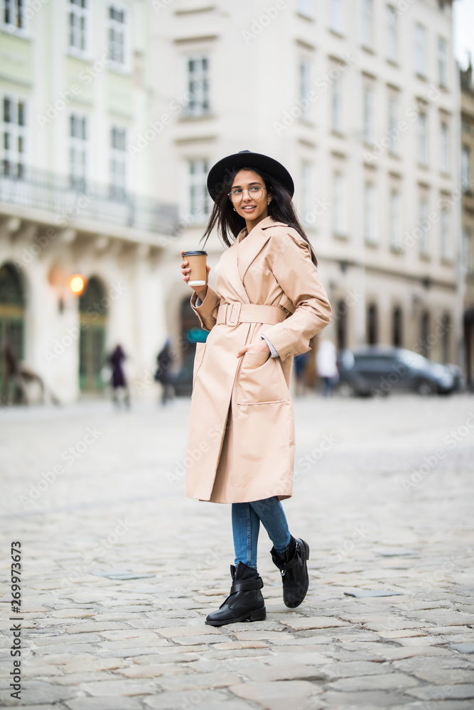 Naklejka premium Young woman wearing coat and black hat walking in the autumn city street and drinking take away coffee in paper cup. Breakfast to go.