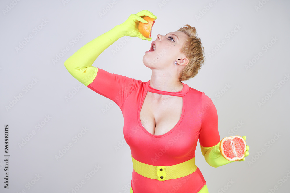 alternative caucasian girl with short hair dressed in sporty red spandex  bodysuit and bright tights with green neon gloves. curvaceous woman holds a  sliced grapefruit on a white studio background Stock Photo