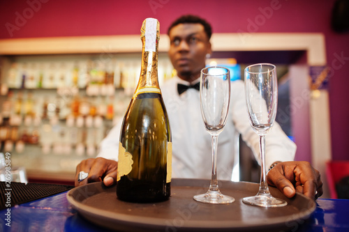 African american bartender at bar holding champagne with glasses on tray.