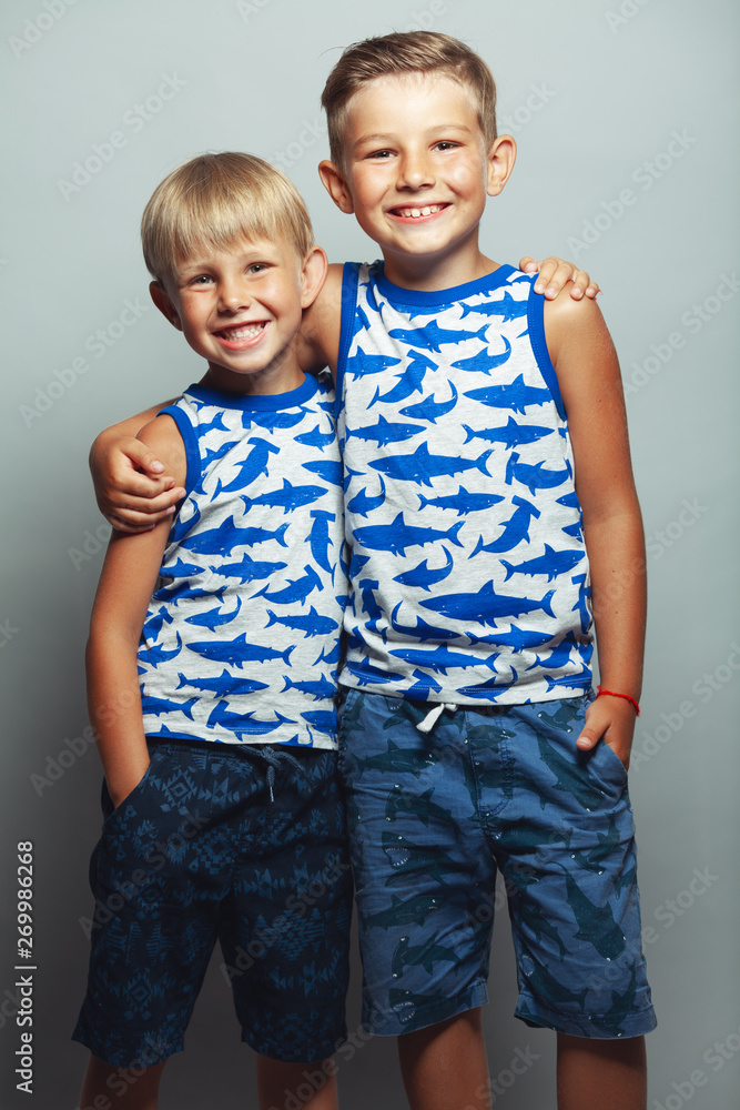 Cute little boys in white and blue sleeveless shirts posing in front of gray background. Portrait of fashionable male childs. Smiling kids posing, hugging. Concept of children style and fashion Cute little boys in white and blue sleeveless shirts posing in front of gray background. Portrait of fashionable male childs. Smiling kids posing, hugging. Concept of children style and fashion
