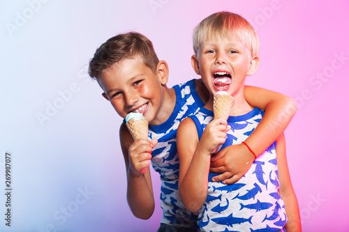 Tasty summer obsession concept. Happy young handsome hipster boys wearing sleeveless shirts with sharks  hugging  eating melting ice cream in waffle cone over pink   blue background. Copy-space