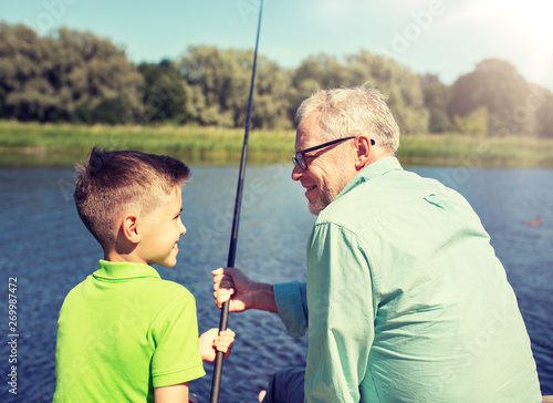 family, generation, summer holidays and people concept - happy grandfather and grandson with fishing rods on river berth