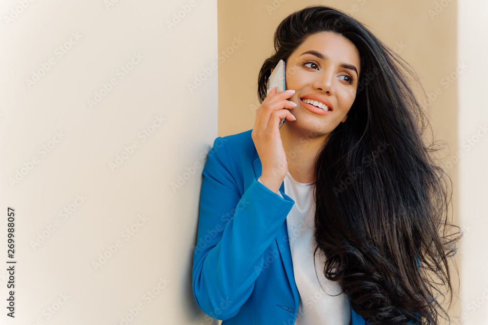Optimistic female entrepreneur with dark wavy hair, wears makeup, smiles happily, holds modern cell phone near ear has conversation dressed formally looks aside, discusses good news in business sphere Optimistic female entrepreneur with dark wavy hair, wears makeup, smiles happily, holds modern cell phone near ear has conversation dressed formally looks aside, discusses good news in business sphere
