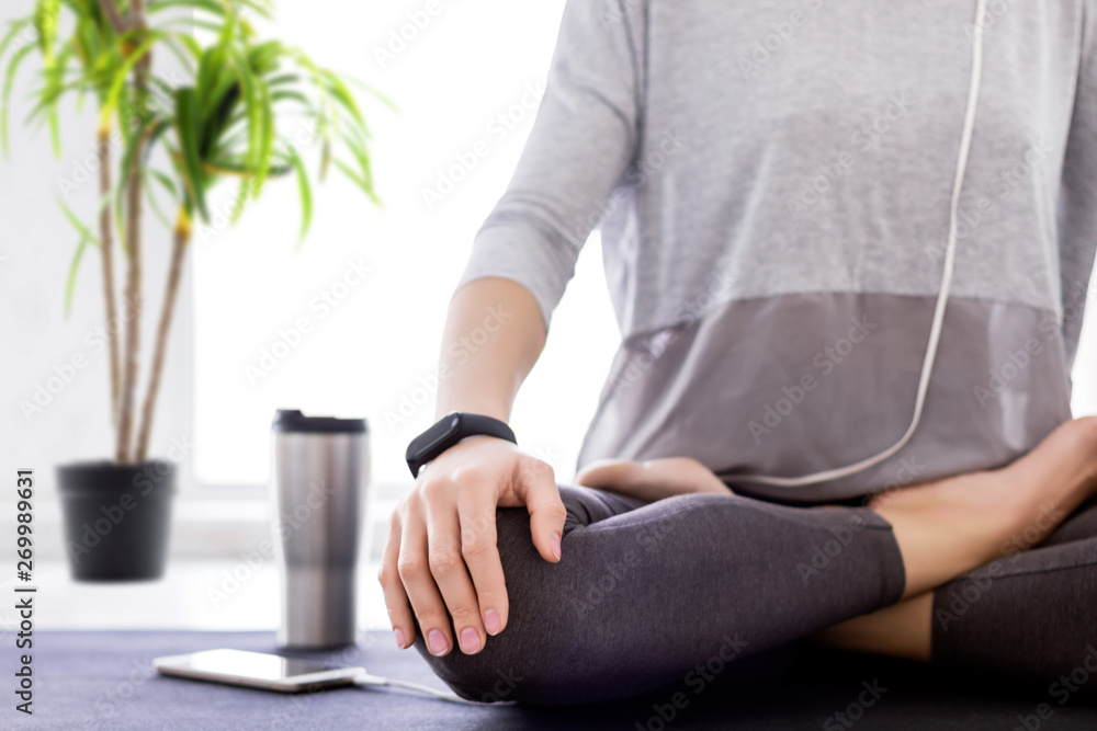 Fototapeta premium Closeup of slender female legs folded crossed in lotus position. Young athletic woman in grey clothes and fitness tracker is practicing yoga, meditating, relaxing while sitting in padma asana at home.