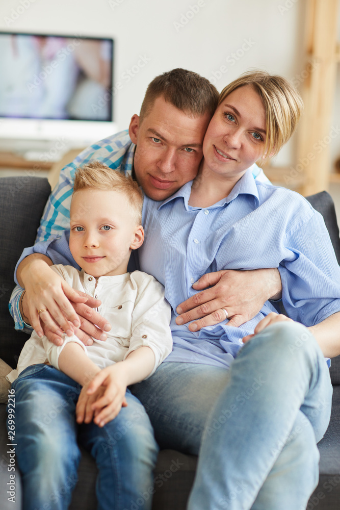 Portrait of positive loving young family sitting on sofa in living room, handsome father hugging wife and son
