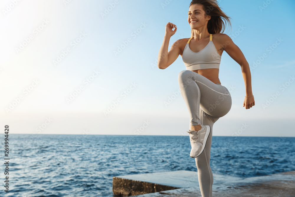 Motivated happy sporty woman wearing sports bra, sneakers enjoying  excercise, training outdoors near sea, workout quay, jogging, running,  jumping energized, smiling during productive fitness session Stock Photo