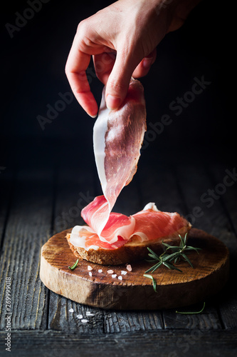 Female hand putting prosciutto on bread over dark wooden background. Sandwich cooking. photo