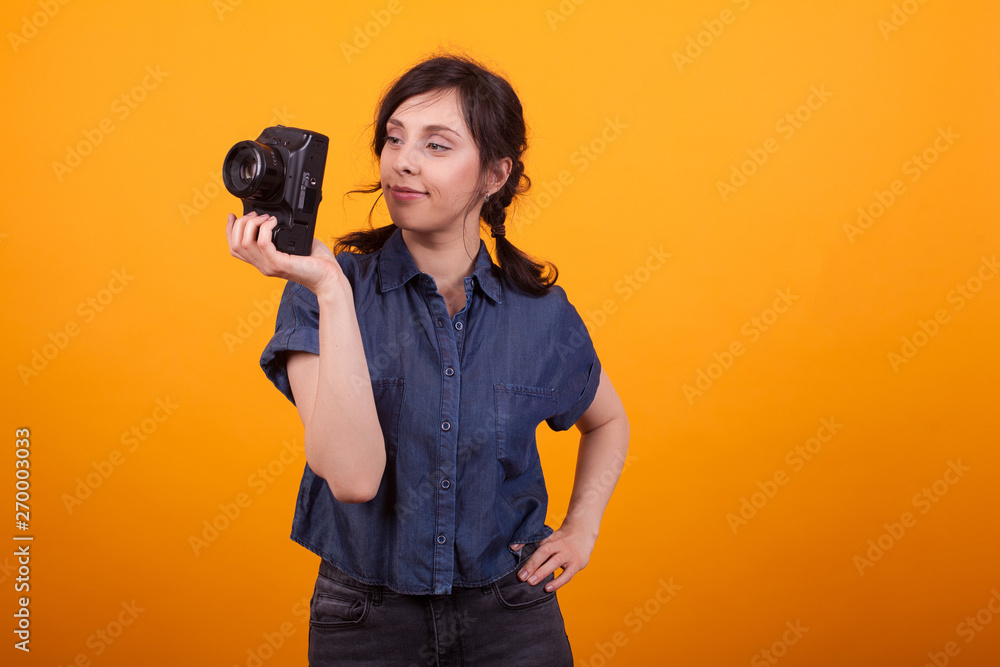 Fototapeta premium Portrait of young female photographer holding her camera in studio over yellow background