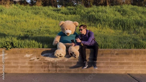 Caucasian guy leans into teddy bear friend, pointing at phone screen, explaining concept matter of factly, energetically. Trying to help teddy. Slow motion. photo