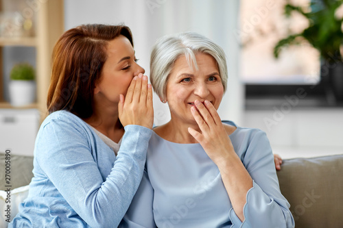 family, generation and people concept - adult daughter whispering secret to senior mother at home