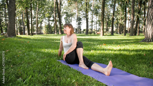 Image of smiling middle aged woman in fitness clothes doing stretching and yoga exercises. WOman meditating and doings sports on fitness mat on grass at park