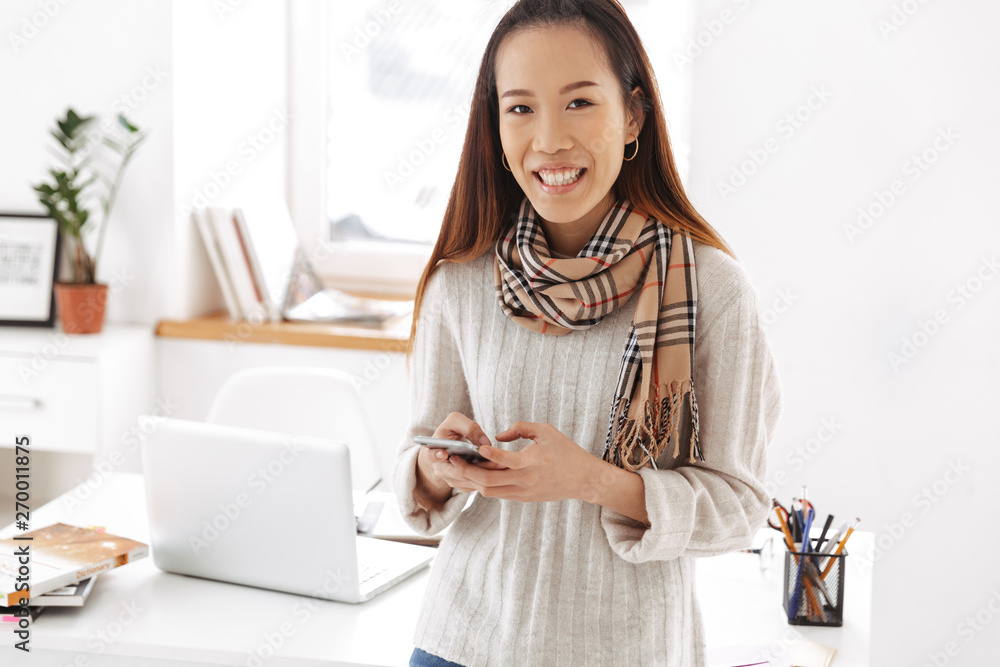Photo of optimistic asian female worker using cellphone and smiling while sitting on table in office