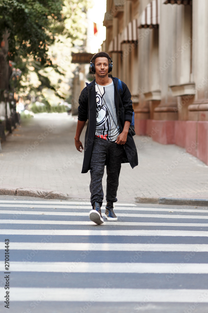 Hipster guy walking with atitudine on the crosswalk listening a music, goes out to work in day time, promotes walking. Outside shot. Hipster guy walking with atitudine on the crosswalk listening a music, goes out to work in day time, promotes walking. Outside shot.