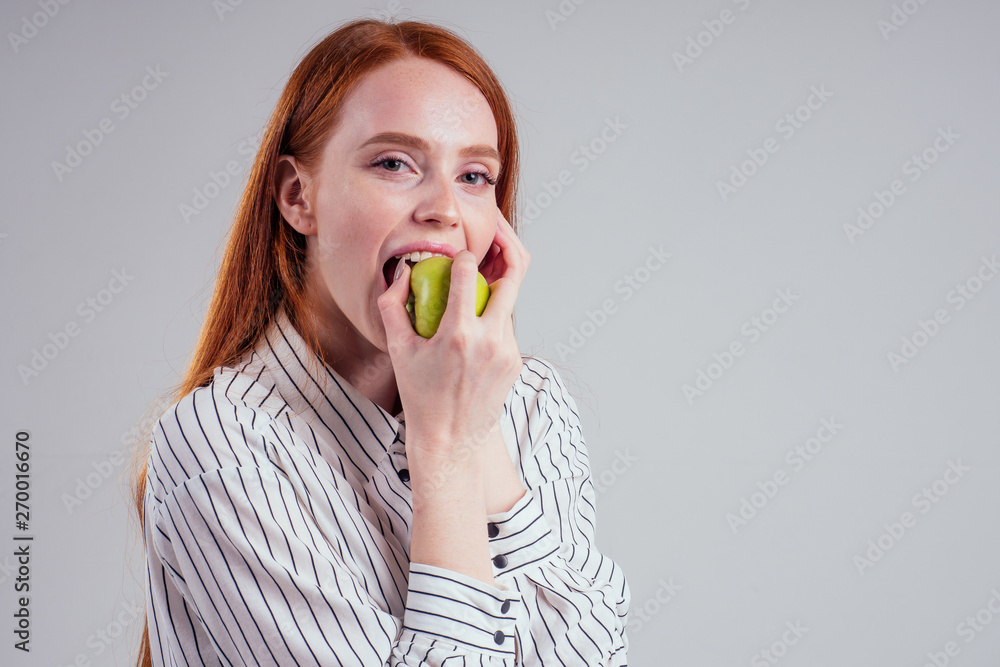 beautiful young redhead businesswoman in a striped shirt snow-white smile and perfect skin eating detox green apple in studio white background(healthy snack student) beautiful young redhead businesswoman in a striped shirt snow-white smile and perfect skin eating detox green apple in studio white background(healthy snack student)